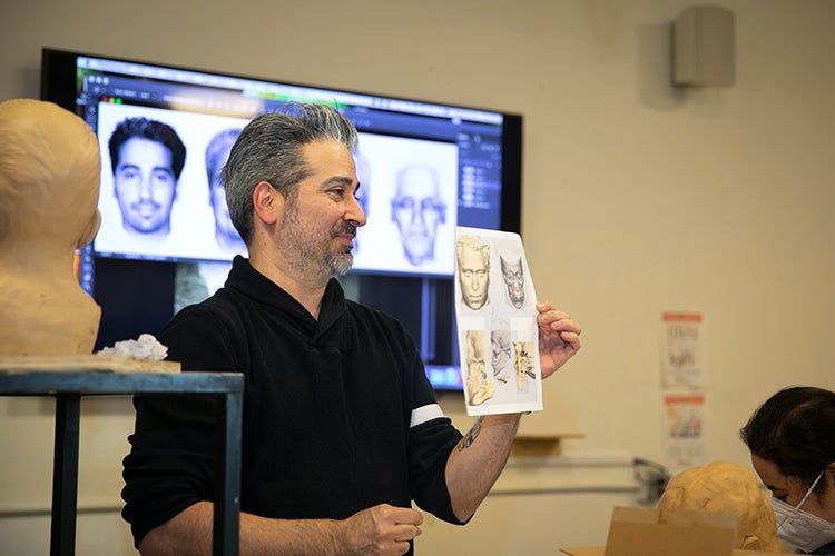 Joe Mullins stands in front of the sculpture class facing the students with a screen behind him showing facial reconstruction techniques