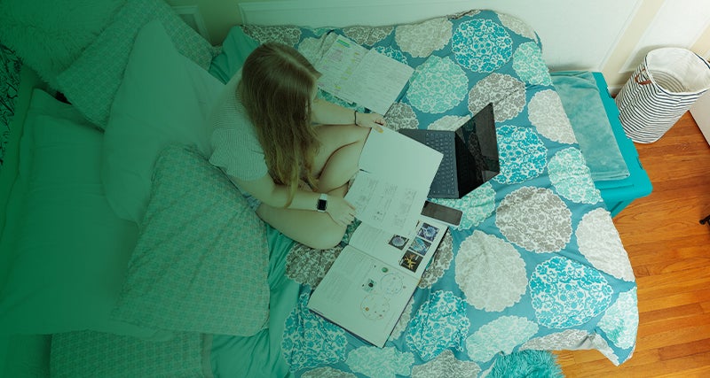 A young person sits on a bed looking at a textbook and notes 