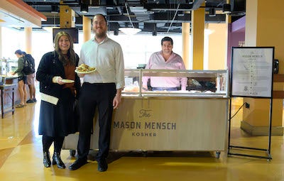 Two people stand in front of a food stand inside Southside dining hall. Another person, looking like they are prepared to serve food, stands behind the card.