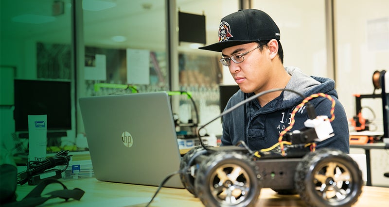 A Mason student sits at a table using a laptop while surrounded by technological equipment 
