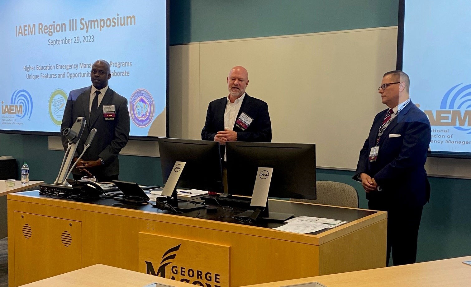 Three men wearing nametags stand at the front of a room, behind a presenter computer setup. Behind them is a screen projecting their powerpoint.