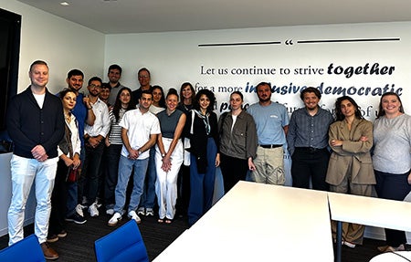 A group of people standing together for a picture in front of a wall mural reading "let us continue to strive together for a more inclusive, democratic, and peaceful" with the rest of the text obscured