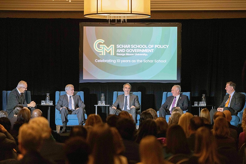 Wide shot of the full stage with five men seated in chairs and an audience in the foreground.