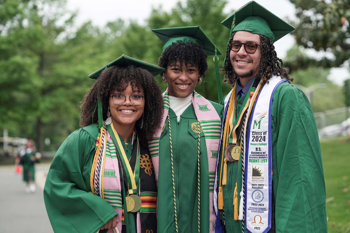 Three graduates stand together and smile on campus