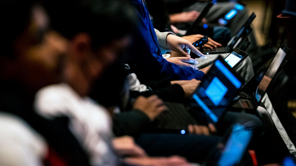 People sit in a row of auditorium seating. Their faces are blurred as the camera focusses on their hands, all using laptops or smartphones.