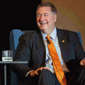 Former Virginia Governor George Allen sits in a blue armchair on stage, smiling slightly, wearing a dark suit with an orange tie.