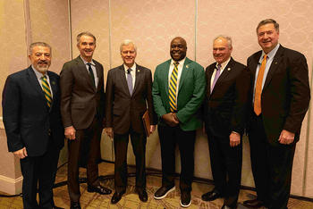 Six men pose shoulder-to-shoulder for a group photo in a hotel meeting space, including four former Virginia governors with David Ramadan and George Mason University President Gregory Washington at center, all in suits and business attire.