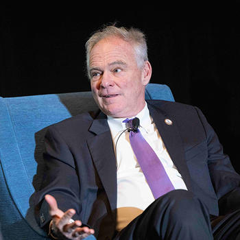 Governor Tim Kaine sits relaxed in a blue armchair on stage, smiling, wearing a dark suit and light-colored tie, with a glass of water on a nearby table.